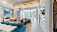 This contemporary single-floor community clubhouse or leasing center is designed with a bold, resort-inspired aesthetic that immediately captures attention. The grand entrance foyer sets an impressive tone with soaring double-height ceilings, warm wood-paneled accent walls, and an eye-catching sculptural ceiling installation with cascading globe pendant lighting. Large-format tile flooring flows throughout, lending a clean, polished continuity to the space.

The main lounge area is a focal point of the facility, featuring vibrant teal velvet sofas, layered area rugs, and a sleek linear fireplace set into a horizontally-striped textured accent wall. A striking geometric chandelier hangs above, adding an artistic dimension to the room. Floor-to-ceiling glass walls and wide glass doors frame panoramic views of the outdoor pool and palm-tree-lined surroundings, bathing the space in abundant natural light.

Beyond the lounge, a hallway with architectural arched lighting details leads to additional rooms, including a dining and meeting area furnished with modern tables and chairs beneath a teal-toned accent wall. A separate bright room with large windows offers additional seating, suitable for work or social gatherings.

Throughout, the design balances vibrant color accents with neutral tones, creating an environment that is both energetic and welcoming — ideal for community gathering and daily use.