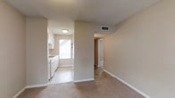 This single-story space presents a straightforward, functional layout with an open feel connecting the main living area to the kitchen. The living space is finished with wall-to-wall carpeting in a neutral beige tone, while the kitchen and adjacent entry area transition to a hard surface flooring. The kitchen is equipped with white cabinetry, a dishwasher, and counter space along one wall, with a window near the entry that allows natural light to filter into the area. Walls throughout are painted in a consistent warm beige, contributing to a cohesive and calm interior palette. A textured ceiling runs throughout the space, and HVAC vents are visibly integrated into both the ceiling and upper wall areas, indicating a central climate control system. A structural column or partial wall separates the living area from the kitchen and entry corridor, creating subtle definition between the spaces without fully closing them off. A hallway extends from the main living area, leading toward additional rooms at the rear of the unit. The overall ambiance is clean, neutral, and move-in ready, offering a blank canvas for personalization.
