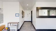 Located at 951 S Oxford Ave in Los Angeles, this building hallway serves as the primary entry and common circulation space for residents. The layout is composed of two connected areas: a front lobby entry and an extended corridor leading to interior building access points.

The entry area features large frosted glass sliding doors that allow diffused natural light to filter into the space while maintaining privacy. A prominent bank of individual metal mailboxes lines one wall, providing organized mail and package management for all residents. A small utility table nearby offers a convenient surface for sorting deliveries.

The corridor extends deeper into the building, finished with large-format gray tile flooring and crisp white walls accented by dark baseboards. Recessed ceiling lighting provides even, functional illumination throughout. A bold graphic wall-mounted artwork adds a decorative focal point along the hallway.

At the rear of the space, a black-framed pass-through window is built into the wall, alongside a secured door providing access to the garage or utility areas. A compact seating area with a modern black leather chair is positioned nearby, offering a brief waiting spot for residents or guests. Security cameras are visibly installed, and clear building notices are posted throughout, reflecting a well-managed residential environment.