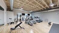 Located at 20300 Civic Center Dr in Southfield, Michigan, this fitness center offers a spacious, single-floor exercise room designed to support a full range of workout routines. The open, exposed truss ceiling lends the space an industrial character while contributing to an airy, high-ceiling feel that enhances the sense of openness throughout.

The main exercise area features light hardwood flooring that extends across much of the space, complemented by rubber matting in designated strength and free-weight zones. The cardio section is well-appointed with multiple elliptical trainers, treadmills, a recumbent bike, and a rowing machine, all arranged to maximize floor space and usability. Wall-mounted flat-screen TVs are positioned throughout for entertainment during workouts.

A dedicated strength area includes a power rack with a barbell and weight plates, TRX suspension straps, and a well-stocked dumbbell rack covering a range of weights. Additional equipment includes a cable weight machine and an adjustable workout bench, rounding out the facility's strength training offerings.

Natural light filters in through multiple windows fitted with roller shades, brightening the gray-toned walls and giving the space a clean, modern aesthetic. A storage unit near the entrance holds yoga mats, exercise balls, and resistance bands, ensuring a variety of fitness options are readily accessible. Full-wall mirrors further enhance the sense of space and allow for form monitoring during training sessions.