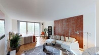 This single-floor space offers an open, airy layout that combines living and sleeping areas in one cohesive environment. The most prominent architectural feature is a large, textured rust-orange accent wall that anchors the living area, providing a bold contrast against the clean white walls and smooth ceiling throughout the space. Natural light floods the interior through expansive floor-to-ceiling sliding glass doors that open to an outdoor balcony, offering views of lush greenery beyond. The flooring is a warm, checkered parquet wood pattern that adds a classic and refined character to the space. The living area is centered around a light-toned sectional sofa paired with a modern rectangular coffee table and a patterned area rug, creating a comfortable and stylish gathering zone. Adjacent to the living space, the sleeping area is defined by a bed dressed in warm, earthy tones that complement the overall color palette. A slim black display cabinet and a potted indoor plant add decorative depth near the balcony entrance, while a reading chair completes the corner seating arrangement. The overall ambiance is warm, modern, and welcoming, with a thoughtful balance of natural materials and contemporary furnishings.