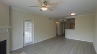 This single-floor residence offers an open and airy layout designed for comfortable everyday living. The main living area is finished with smooth, modern laminate flooring in a neutral gray tone that extends throughout the space, creating a clean and cohesive aesthetic. A classic ceiling fan with an integrated light fixture is centrally mounted, providing both airflow and ambient lighting to the room.

Natural light enters through a windowed door fitted with horizontal blinds, offering both brightness and privacy. The warm, neutral-toned walls complement the flooring and contribute to an inviting atmosphere throughout.

The living area flows seamlessly into a connected kitchen, which is partially visible beyond a raised breakfast bar counter. The kitchen features warm wood-toned cabinetry and appears to include standard appliances, making it a practical and efficient space. The bar-height counter serves as a natural divider between the living and kitchen areas while maintaining the open feel of the floor plan.

A hallway visible in the background suggests additional rooms beyond the main living space. Ceiling-mounted flush lights provide supplemental illumination throughout the unit. The overall design is straightforward and functional, with consistent finishes that create a unified, move-in-ready environment.