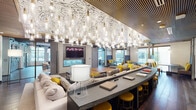 This contemporary community lounge is a thoughtfully designed gathering space that blends striking aesthetics with functional versatility. The centerpiece of the main lounge is a breathtaking installation of cascading glass pendant lights suspended from an artfully patterned ceiling, creating an atmosphere that is both dramatic and inviting. Slatted wood ceiling panels add warmth and texture, complementing the rich hardwood flooring that flows throughout the space.

The lounge is organized into distinct zones to accommodate a variety of uses. A generous seating area features plush sofas and chairs accented with bold yellow throw pillows, arranged around a long, curved bar-height counter with matching yellow stools — ideal for socializing or working. A dedicated booth area lines the perimeter, offering semi-private high-backed seating with individual tables and direct views of a lushly landscaped outdoor area through floor-to-ceiling windows that flood the interior with natural light.

A fully equipped kitchenette, finished with subway tile backsplash, open metal shelving, dark countertops, and stainless steel appliances, provides convenient amenities for residents or guests. The entry transition zone features large-format stone tile flooring, exposed ductwork, and a wall-mounted display, lending an industrial-modern character. Glass-paneled sliding doors frame views into the lounge, connecting the spaces with transparency and openness throughout.