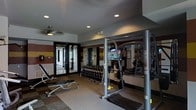 This fitness center is a thoughtfully designed, single-floor facility built to accommodate a wide variety of exercise routines. The space features a practical and functional layout, with recessed lighting and a ceiling fan overhead that help maintain a comfortable workout environment.

A standout feature of the gym is the full wall of mirrors, which enhances the sense of space while allowing users to monitor their form during exercise. The walls are accented with bold horizontal color-block stripes in deep red and gold tones, adding a dynamic visual character to the room.

The facility is well-stocked with strength training equipment, including a multi-functional cable and weight machine, a dumbbell rack with a comprehensive range of free weights, and an adjustable weight bench. A flat-screen television is mounted on the mirrored wall, offering entertainment during workouts. The flooring features a combination of carpet and rubberized mat surfaces, providing both comfort and safety.

Large glass double doors open to an adjacent corridor, allowing natural light to filter into the space and providing easy access. Overall, the fitness center is a clean, organized, and fully equipped environment designed to meet the needs of a broad range of fitness levels and training goals.