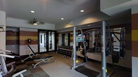 This fitness center is a thoughtfully designed, single-floor facility built to accommodate a wide variety of exercise routines. The space features a practical and functional layout, with recessed lighting and a ceiling fan overhead that help maintain a comfortable workout environment.

A standout feature of the gym is the full wall of mirrors, which enhances the sense of space while allowing users to monitor their form during exercise. The walls are accented with bold horizontal color-block stripes in deep red and gold tones, adding a dynamic visual character to the room.

The facility is well-stocked with strength training equipment, including a multi-functional cable and weight machine, a dumbbell rack with a comprehensive range of free weights, and an adjustable weight bench. A flat-screen television is mounted on the mirrored wall, offering entertainment during workouts. The flooring features a combination of carpet and rubberized mat surfaces, providing both comfort and safety.

Large glass double doors open to an adjacent corridor, allowing natural light to filter into the space and providing easy access. Overall, the fitness center is a clean, organized, and fully equipped environment designed to meet the needs of a broad range of fitness levels and training goals.