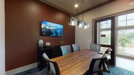 This well-appointed conference room offers a refined and functional setting for meetings and collaborative work. The room centers around a striking live-edge wood conference table, which seats several guests in a mix of upholstered and ergonomic task chairs, providing both comfort and practicality. A wall-mounted flat-screen display is positioned on a rich, deep-red accent wall, accompanied by a dark credenza adorned with decorative vessels — adding a sophisticated, curated touch to the space. Overhead, an industrial-style linear pendant light fixture with glass shades casts warm, focused illumination across the table. The room features a patterned blue-toned carpet that anchors the space with depth and texture. Tall, dark-framed glass doors open to an adjacent common area, flooding the room with natural light and offering views of lush greenery outside. A large window along one wall further enhances the connection to the outdoors. Vibrant abstract artwork displayed near the entry adds a creative energy. The overall design balances a warm, contemporary aesthetic with professional functionality, making it an inviting and well-equipped space for focused gatherings.