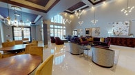 This elegantly appointed single-floor amenity space exudes luxury and refinement at every turn. The grand main lounge commands attention with soaring double-height ceilings, expansive floor-to-ceiling windows that flood the room with natural light, and dramatic multi-globe chandelier fixtures. Rich jewel-toned furnishings — deep plum sofas, gold velvet chairs, and patterned accent pieces — are arranged across gleaming large-format white tile floors, creating a welcoming yet upscale atmosphere.

Adjacent seating areas provide intimate gathering spaces, including a cozy lounge with a wall-mounted television, vibrant abstract artwork, and tailored tufted seating. A separate dining area features warm wood round tables, gold velvet chairs, and stylish pendant lighting beneath coffered ceilings accented in deep burgundy tones.

The space includes two wet bar stations — one highlighted by an ornate onyx stone surround and matching countertops, and another featuring a tufted upholstered accent wall with marble countertops and dark cabinetry. A fully equipped catering kitchen with granite countertops, dark wood cabinets, and stainless steel appliances supports event hosting needs.

Bold black-framed glass doors define transitions between zones, while decorative columns add architectural character throughout. The cohesive design palette of burgundy, gold, and neutral tones lends the entire space a polished, upscale identity.