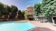 This inviting outdoor amenity space is centered around a generously sized, freeform swimming pool with clear turquoise water, set within a broad, terracotta-toned concrete sun deck. The pool area is thoughtfully appointed with lounge seating and shaded umbrella stations, offering residents a comfortable place to relax and unwind. Adjacent to the main pool, a circular spa provides a dedicated space for relaxation, surrounded by tasteful railings and complemented by additional seating. The courtyard is enclosed by multi-story residential buildings featuring warm tan stucco exteriors, private balconies, and large windows that overlook the amenity area. Mature palm trees and a rich assortment of tropical and subtropical landscaping, including tall palms, leafy trees, and ornamental shrubs, lend a resort-like atmosphere to the setting. Iron fencing borders the pool and spa areas, providing a sense of security and structure while maintaining an open, airy feel. The overall design creates a cohesive, well-maintained outdoor retreat that balances functionality with aesthetic appeal, offering an attractive communal gathering space bathed in natural sunlight throughout the day.