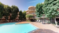 This inviting outdoor amenity space is centered around a generously sized, freeform swimming pool with clear turquoise water, set within a broad, terracotta-toned concrete sun deck. The pool area is thoughtfully appointed with lounge seating and shaded umbrella stations, offering residents a comfortable place to relax and unwind. Adjacent to the main pool, a circular spa provides a dedicated space for relaxation, surrounded by tasteful railings and complemented by additional seating. The courtyard is enclosed by multi-story residential buildings featuring warm tan stucco exteriors, private balconies, and large windows that overlook the amenity area. Mature palm trees and a rich assortment of tropical and subtropical landscaping, including tall palms, leafy trees, and ornamental shrubs, lend a resort-like atmosphere to the setting. Iron fencing borders the pool and spa areas, providing a sense of security and structure while maintaining an open, airy feel. The overall design creates a cohesive, well-maintained outdoor retreat that balances functionality with aesthetic appeal, offering an attractive communal gathering space bathed in natural sunlight throughout the day.