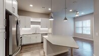 This single-floor unit showcases a clean, contemporary design throughout. The open-concept kitchen and living area serves as the heart of the space, anchored by a generously sized central island with a white quartz countertop and seating. The kitchen is outfitted with white shaker-style cabinetry, a white subway tile backsplash with under-cabinet lighting, and a full suite of stainless steel appliances, including a refrigerator, range, microwave, and dishwasher. Two industrial-style pendant lights hang above the island, complementing the recessed lighting overhead. Wood-look plank flooring runs throughout the unit, providing a warm, cohesive aesthetic that contrasts with the neutral gray-toned walls and crisp white trim. The living area receives natural light through window blinds and is served by a ceiling fan. A hallway connects the main living space to the private rooms, where a bedroom offers a window with natural light and easy access to adjacent spaces. The bathroom features a tiled tub-shower combination with a curved curtain rod and wood-look flooring. A stacked washer and dryer unit is conveniently housed within the unit, adding practical functionality to the overall well-appointed layout.