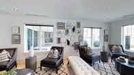 This single-story space showcases a thoughtfully designed interior with a cohesive contemporary aesthetic throughout. The lounge area serves as a focal point, featuring light hardwood flooring, large windows on multiple walls, and an eye-catching gallery wall arrangement of mixed-media framed artwork in a monochromatic palette. Dark upholstered accent chairs paired with patterned throw pillows, a tufted bench, and a black-and-white geometric area rug contribute to the sophisticated, layered look. Multiple windows and sliding glass doors flood the space with natural light while offering views of surrounding outdoor areas with lush greenery.

The reception or entry area transitions seamlessly from the lounge, featuring striking herringbone-patterned parquet flooring, black-framed glass partition doors, and recessed lighting that enhances the bright, open atmosphere. A separate workspace or office area includes soaring ceilings clad in dark corrugated metal paneling, a prominent sliding chevron-patterned barn door, a pendant light fixture, and steel-framed glass partitions. Together, these spaces deliver a well-balanced blend of industrial and modern design elements, creating an inviting and functional environment throughout.
