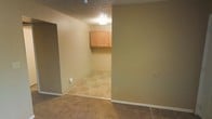 This single-floor space is laid out with a clear distinction between its functional zones. The main area features wall-to-wall carpet in a neutral tone, providing a comfortable and practical surface underfoot. Smooth, beige-painted walls run throughout, complemented by white baseboards that offer a clean, finished appearance. A textured popcorn ceiling adds a classic touch overhead, with a flush-mount light fixture providing basic illumination.

An open transition leads into a secondary room finished with patterned tile flooring in warm earth tones, suggesting its use as a utility or laundry area. This room features upper wall-mounted cabinetry in light natural wood, offering practical storage space. The tile flooring in this area is both durable and easy to maintain, well-suited for a utility-focused purpose. An electrical outlet is visible along the wall, supporting appliance hookups. The overall palette throughout the space is understated and cohesive, with tan and beige tones creating a calm, neutral ambiance. The layout is straightforward and functional, making efficient use of the available square footage across its single level.