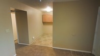 This single-floor space is laid out with a clear distinction between its functional zones. The main area features wall-to-wall carpet in a neutral tone, providing a comfortable and practical surface underfoot. Smooth, beige-painted walls run throughout, complemented by white baseboards that offer a clean, finished appearance. A textured popcorn ceiling adds a classic touch overhead, with a flush-mount light fixture providing basic illumination.

An open transition leads into a secondary room finished with patterned tile flooring in warm earth tones, suggesting its use as a utility or laundry area. This room features upper wall-mounted cabinetry in light natural wood, offering practical storage space. The tile flooring in this area is both durable and easy to maintain, well-suited for a utility-focused purpose. An electrical outlet is visible along the wall, supporting appliance hookups. The overall palette throughout the space is understated and cohesive, with tan and beige tones creating a calm, neutral ambiance. The layout is straightforward and functional, making efficient use of the available square footage across its single level.