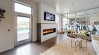 This single-story clubhouse is designed with a contemporary aesthetic, offering a well-appointed collection of shared amenity spaces. The main lounge area serves as a focal point, featuring a sleek, wall-mounted linear fireplace flanked by a flat-screen TV above. Warm wood-tone flooring runs throughout, complemented by plush area rugs and curated décor accents in gold tones. A glass partition wall separates the lounge from an adjacent fitness area, which is visible through the transparent divide and equipped with exercise machines. Direct access to an outdoor pool area is provided through a glass door in the lounge.

The kitchen area is outfitted with rich warm-wood cabinetry, white quartz countertops, a professional-style pull-down faucet, an undermount sink, a beverage refrigerator, and a dishwasher. A continuous bank of large, grid-pane windows lines the kitchen wall, flooding the space with natural light and offering views of the surrounding area and glimpses of water beyond.

The lobby presents a clean, open design with floor-to-ceiling glass partition walls, tiled flooring, and teal upholstered ottomans paired with a metallic accent table, creating an inviting and polished entrance to the clubhouse.