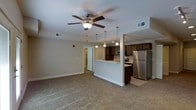 This single-floor residence offers a well-designed open-concept layout that seamlessly connects the living area to the kitchen. The spacious living room features plush wall-to-wall carpeting and a ceiling fan with a light fixture, creating a comfortable and functional environment. Neutral-toned walls and clean white baseboards contribute to a bright, airy atmosphere throughout the space.

The kitchen is defined by a raised peninsula counter that serves as a natural divider between the cooking and living areas. Dark-toned cabinetry lines the kitchen walls, providing ample storage, while pendant lights hang above the peninsula to create focused task lighting. Stainless steel appliances, including a full-size refrigerator and range, lend a modern touch to the kitchen. Tile flooring in the kitchen area contrasts with the carpeted living space, clearly delineating each zone.

A hallway visible from the main living area leads to additional rooms, suggesting a practical and efficient floor plan. HVAC vents are integrated into the ceiling throughout, ensuring year-round climate comfort. Overall, the space presents a clean, functional, and inviting interior suited for everyday living.