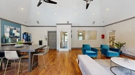 This well-appointed single-floor communal space makes a strong first impression with its dramatic vaulted ceilings that lend an open, airy feel throughout. Rich hardwood flooring runs seamlessly across the entire space, grounding the bright, neutral-toned walls accented by crisp gray crown molding. Dual ceiling fans keep the atmosphere comfortable while adding a refined architectural touch.

The space is thoughtfully divided into functional zones. A lounge area features plush teal velvet armchairs arranged around a marble-top coffee table, set atop a soft area rug, offering a relaxed setting for conversation or informal gatherings. Adjacent to it, a full-length sofa and bench seat face a glass patio door, which floods the area with natural light and provides direct outdoor access.

A dining and collaboration area anchors the opposite end of the space, centered around a substantial dark wood high-top table surrounded by modern gold-accented bar chairs. Patterned drapery frames large windows that invite ample natural light. A teal accent wall with decorative artwork and a credenza with a beverage station adds a pop of color and practical amenity. A pass-through window connects to an adjacent office or service area, suggesting a leasing or management function. The overall ambiance is modern, welcoming, and purposefully designed for community use.