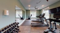 This fitness center is a thoughtfully designed, single-room workout facility that offers a comprehensive range of exercise equipment in a welcoming environment. The room is finished with soft sage-green walls and durable carpet tile flooring, creating a clean and energizing atmosphere. Recessed lighting and a ceiling fan ensure the space remains well-lit and comfortable during workouts.

Natural light fills the room through multiple windows fitted with blinds, offering views of the surrounding neighborhood. A large wall-mounted mirror spans a significant portion of one wall, visually expanding the space and allowing users to monitor their form during exercise.

The facility is outfitted with multiple True-brand treadmills and a stair climber, providing a variety of cardio options. An Echelon connected spin bike with a digital display screen adds a modern, interactive training element. A wall-mounted flat-screen television is positioned for easy viewing from the cardio equipment. A dumbbell rack stocked with a range of free weights supports strength training, while a padded exercise mat on the floor accommodates stretching and floor-based workouts.

Overall, this fitness center balances functionality and comfort, delivering a well-rounded workout experience within a bright, neatly maintained space.