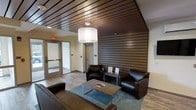 This single-floor space makes a strong first impression with its thoughtfully designed lounge area, combining modern aesthetics with a comfortable, professional atmosphere. The seating area is anchored by rich, dark leather armchairs and a sofa arranged around a rustic wood-and-metal coffee table, set atop a teal area rug that adds a subtle pop of color to the warm interior palette. A striking feature of the space is the dark wood slat wall panel that serves as a bold architectural backdrop, complementing the matching slatted wood ceiling detail that adds depth and visual interest overhead. A large pendant drum light fixture hangs from the ceiling, providing warm, ambient illumination throughout the lounge. The flooring is finished in wood-look planks that carry the cohesive warm-toned design throughout. Glass-paneled entry doors allow natural light to filter into the space, enhancing its open and inviting feel. The overall design balances function and style, making it well-suited as a welcoming reception or common lounge area. Informational materials are displayed on a side table, suggesting the space is designed to serve and inform visitors upon arrival.