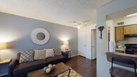 This single-floor residence offers a thoughtfully designed layout that balances comfort and functionality across its key living areas. The living room serves as the heart of the home, featuring rich dark hardwood-style flooring, a textured ceiling, and a ceiling fan overhead. Cool gray accent walls complement the neutral tones throughout, and a large window dressed with patterned sheer curtains allows natural light to filter in, offering a pleasant view of the greenery outside. The space comfortably accommodates a seating arrangement anchored by a coffee table and area rug, creating a welcoming gathering area. The galley-style kitchen is lined on both sides with warm wood cabinetry and equipped with black appliances, including a range, refrigerator, and microwave, alongside ample counter space and a double sink — providing everything needed for everyday cooking. The bedroom is a serene retreat finished with plush carpeting, soft white walls, and a textured ceiling. A window dressed with decorative sheer curtains brings in soft natural light, while generous space accommodates a full dresser and bedside tables. The room's neutral tones and understated finishes create a calm, restful ambiance throughout.