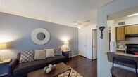 This single-floor residence offers a thoughtfully designed layout that balances comfort and functionality across its key living areas. The living room serves as the heart of the home, featuring rich dark hardwood-style flooring, a textured ceiling, and a ceiling fan overhead. Cool gray accent walls complement the neutral tones throughout, and a large window dressed with patterned sheer curtains allows natural light to filter in, offering a pleasant view of the greenery outside. The space comfortably accommodates a seating arrangement anchored by a coffee table and area rug, creating a welcoming gathering area. The galley-style kitchen is lined on both sides with warm wood cabinetry and equipped with black appliances, including a range, refrigerator, and microwave, alongside ample counter space and a double sink — providing everything needed for everyday cooking. The bedroom is a serene retreat finished with plush carpeting, soft white walls, and a textured ceiling. A window dressed with decorative sheer curtains brings in soft natural light, while generous space accommodates a full dresser and bedside tables. The room's neutral tones and understated finishes create a calm, restful ambiance throughout.