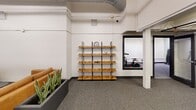 Located at 1110 Washington Ave in downtown St. Louis, this single-floor amenity space offers a well-rounded combination of social and fitness facilities. The layout is anchored by an open lounge area furnished with tan leather sofas arranged over a colorful area rug, complemented by a wooden table and chairs. A freestanding open shelving unit with metal framing and warm wood shelves adds a modern decorative element, while potted greenery introduces a touch of life to the space. Adjacent to the lounge, a wall-mounted mailbox bank and glass-paneled doors provide functional building amenity access.

The fitness center occupies a dedicated area with noticeably elevated ceilings, exposed ductwork, and track lighting. It is equipped with a treadmill, stationary spin bike, a full dumbbell rack, a TRX suspension training station, a weight bench, and a smart interactive mirror for guided workouts. Full-length mirrors line the fitness area walls, enhancing the sense of openness. Large interior windows connect the gym visually to the lounge area, maintaining a sense of connectivity throughout the floor. Neutral-toned carpet tiles, white walls, and industrial-style exposed mechanical systems give the space a clean, modern aesthetic consistent with an urban residential building's amenity offering.