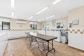 Located at 1 Albany St in Fort Erie, Ontario, this communal laundry facility is a practical and well-organized space designed to serve its users efficiently. The room is bright and welcoming, illuminated by a series of fluorescent ceiling fixtures that ensure consistent lighting throughout.

The laundry area is outfitted with a full row of commercial-grade top-loading washing machines and front-loading dryers, all equipped with modern digital payment interfaces for convenient coin-free operation. A dedicated front-loading washer is also available, offering versatility for different laundry needs. Built-in countertop surfaces alongside the machines provide additional workspace for sorting and organizing laundry.

Centrally positioned within the room are two long folding tables on casters, offering ample surface area for folding and sorting garments. The space also includes a seating area with chairs, allowing users to wait comfortably during their laundry cycles.

The walls feature a warm cream tone accented by a decorative tile border running at mid-height, complemented by warm-toned tile flooring that is both durable and easy to maintain. A wall-mounted air conditioning unit ensures year-round comfort. Informational posted notices are displayed throughout the room. A window allows natural light to supplement the interior lighting, adding to the overall brightness of the space.