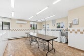 Located at 1 Albany St in Fort Erie, Ontario, this communal laundry facility is a practical and well-organized space designed to serve its users efficiently. The room is bright and welcoming, illuminated by a series of fluorescent ceiling fixtures that ensure consistent lighting throughout.

The laundry area is outfitted with a full row of commercial-grade top-loading washing machines and front-loading dryers, all equipped with modern digital payment interfaces for convenient coin-free operation. A dedicated front-loading washer is also available, offering versatility for different laundry needs. Built-in countertop surfaces alongside the machines provide additional workspace for sorting and organizing laundry.

Centrally positioned within the room are two long folding tables on casters, offering ample surface area for folding and sorting garments. The space also includes a seating area with chairs, allowing users to wait comfortably during their laundry cycles.

The walls feature a warm cream tone accented by a decorative tile border running at mid-height, complemented by warm-toned tile flooring that is both durable and easy to maintain. A wall-mounted air conditioning unit ensures year-round comfort. Informational posted notices are displayed throughout the room. A window allows natural light to supplement the interior lighting, adding to the overall brightness of the space.