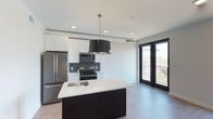 This single-floor, two-bedroom, two-bathroom residence offers a contemporary open-concept layout with clean lines and modern finishes throughout. The kitchen serves as a central highlight, featuring a large dark-based island with a white quartz countertop, an undermount sink, and a striking black drum pendant light overhead. Stainless steel appliances, including a refrigerator, range, and built-in microwave, complement the white shaker-style cabinetry and dark tile backsplash, creating a sharp contrast that defines the space's modern aesthetic. Recessed lighting throughout ensures the area remains bright and functional. Light-toned hardwood-style flooring extends across the open living area, enhancing the sense of spaciousness. A set of floor-to-ceiling black-framed glass doors opens to a private balcony, allowing natural light to flood the interior while offering views of the surrounding neighborhood. The overall ambiance is clean, urban, and well-appointed, making it a functional yet stylish living space suited for contemporary lifestyles.