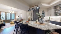 This thoughtfully designed single-floor residence combines modern aesthetics with functional living. The open-concept kitchen is a standout, featuring two-tone cabinetry in light gray and navy, granite countertops, a subway tile backsplash, stainless steel appliances, and a generous island with seating for multiple guests — all illuminated by a trio of pendant lights on a brass rail fixture. The kitchen flows seamlessly into a dining area and an inviting living room, where hardwood-style flooring, a bold navy sofa, and a statement chandelier create a curated, contemporary atmosphere. Floor-to-ceiling glass doors in the living area invite abundant natural light and provide access to an outdoor space with views of the surrounding building exterior. Two well-sized bedrooms offer plush carpeting, ceiling fans, and natural window light — each styled with warm, coordinated decor. The primary bathroom features a wide granite-topped vanity with shaker-style cabinetry and gold hardware, while the secondary bathroom mirrors this refined design with tall upper cabinetry and clean tile work. Throughout the space, gray-toned interior doors with black hardware provide a cohesive architectural detail that ties the overall design together.