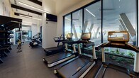 This two-story fitness center is thoughtfully designed to accommodate a comprehensive range of workout activities across both levels. The ground floor serves as the primary cardio and functional training hub, featuring Life Fitness treadmills, ellipticals, and rowing machines positioned along floor-to-ceiling glass walls that frame outdoor views of the surrounding urban landscape. A dedicated spin studio occupies one wing, outfitted with stationary cycling bikes, a wall-mounted display screen, and hard-surface flooring. Bold, colorful fitness-themed murals animate the white walls, adding a dynamic visual character to the space. A staircase with teal metal railings connects to the upper level, with built-in wooden shelving beneath housing rolled towels for convenience. The upper floor houses a well-equipped strength training area, featuring a power rack, cable machines, a full dumbbell rack, benches, medicine balls, and floor-to-ceiling mirrors that visually expand the space. Light wood-style flooring runs throughout both levels, complemented by recessed and linear LED lighting. The overall design balances energetic aesthetics with functional layout, creating an inviting and well-equipped environment for diverse fitness routines.