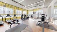 Located at 100 Plyer Cir in Indian Trail, NC, this fitness center offers a generously sized, single-floor workout facility designed to support a full range of exercise activities. The space is defined by warm hardwood-style flooring, elevated coffered ceilings with ceiling fans, and recessed lighting that creates a bright, energizing atmosphere throughout.

Large windows line the walls, flooding the interior with abundant natural light and providing views of the surrounding community. A bold yellow accent wall adorned with motivational fitness typography — featuring words like "dedication," "workout," "training," and "exercise" — adds a dynamic and energizing visual element to the space.

The facility is thoughtfully organized to accommodate multiple workout zones. A cardio section houses a row of treadmills and elliptical machines positioned along the windowed wall. A strength training area features cable machines, a multi-functional rack with resistance bands, kettlebells, and stability balls, complemented by floor mats for stretching or bodyweight exercises. Full-length mirrors are strategically placed to enhance the sense of space and assist with form during workouts. A wall-mounted flat-screen TV provides entertainment options. Double glass doors offer easy access to the exterior, further connecting the space to its surroundings.