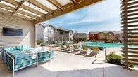 This expansive outdoor amenity space is thoughtfully designed to offer residents a resort-style experience. At its center is a generously sized swimming pool with clear, blue water, complemented by a separate circular hot tub nestled among neatly trimmed shrubbery. The wide pool deck is outfitted with numerous green and yellow lounge chairs, providing ample space for sunbathing and relaxation under the open sky.

A striking wood-framed pergola anchors one end of the space, draped with string lights that add a warm ambiance for evening gatherings. Comfortable striped outdoor seating beneath the pergola offers a shaded retreat. Adjacent to the pergola, a modern slatted wood facade with large glass windows serves as an architectural focal point, lending the area a contemporary aesthetic.

Landscaping throughout is carefully maintained, featuring ornamental shrubs, gravel beds, and young trees that provide greenery without obstructing sightlines. Paved walkways guide movement through the space, connecting the pool area to surrounding community buildings. Black metal security fencing encircles the pool zone for safety, with gated entry access. Adirondack chairs and high-top tables near a raised seating platform offer additional gathering spots. The surrounding multi-story residential buildings frame the space, creating a cohesive and welcoming community environment.