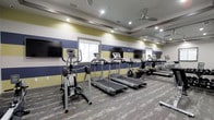 This fitness center offers a clean, purpose-built environment designed to meet a range of cardio and exercise needs. The space is outfitted with multiple treadmills and an elliptical trainer, providing ample options for cardiovascular workouts. Wall-mounted flat-screen televisions are positioned at eye level, allowing users to stay entertained during their sessions. The room's design features a bold striped color scheme of navy blue and gold on the walls, lending the space a dynamic and energetic atmosphere. A full-length mirrored wall on one side visually expands the room, creating a sense of openness while allowing users to monitor their form during exercise. Recessed lighting in the tray ceiling ensures bright, even illumination throughout the space, complemented by a ceiling fan that promotes air circulation for a comfortable workout environment. The flooring consists of durable, patterned carpet suited for a fitness setting. Windows allow natural light to filter in, further enhancing the welcoming ambiance. Overall, this fitness center is thoughtfully designed to balance functionality with an appealing aesthetic, making it an inviting space for daily exercise routines.