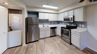 This single-floor space showcases a thoughtfully designed kitchen that serves as a functional and visually appealing centerpiece. The kitchen is outfitted with a full suite of stainless steel appliances, including a refrigerator, electric range with microwave above, and a dishwasher, all complementing the crisp white cabinetry that lines the walls. The cabinetry features raised-panel doors and provides generous storage both above and below the countertops. Light-toned countertops offer ample prep space, while the undermount sink is positioned beneath a dedicated workspace area. The flooring throughout is a warm wood-look laminate or vinyl plank, adding texture and warmth to the otherwise neutral palette. Soft blue-gray accent walls provide subtle contrast against the white cabinetry, contributing to a calm, modern aesthetic. Overhead, a flush-mounted fluorescent light fixture ensures bright, even illumination throughout the kitchen workspace. The overall design balances practicality with a clean, contemporary style, making the kitchen both efficient and inviting for everyday use.