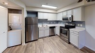 This single-floor space showcases a thoughtfully designed kitchen that serves as a functional and visually appealing centerpiece. The kitchen is outfitted with a full suite of stainless steel appliances, including a refrigerator, electric range with microwave above, and a dishwasher, all complementing the crisp white cabinetry that lines the walls. The cabinetry features raised-panel doors and provides generous storage both above and below the countertops. Light-toned countertops offer ample prep space, while the undermount sink is positioned beneath a dedicated workspace area. The flooring throughout is a warm wood-look laminate or vinyl plank, adding texture and warmth to the otherwise neutral palette. Soft blue-gray accent walls provide subtle contrast against the white cabinetry, contributing to a calm, modern aesthetic. Overhead, a flush-mounted fluorescent light fixture ensures bright, even illumination throughout the kitchen workspace. The overall design balances practicality with a clean, contemporary style, making the kitchen both efficient and inviting for everyday use.