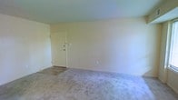 This single-floor space offers a straightforward and functional layout designed for everyday living. The interior is characterized by neutral, light-toned walls that create a calm and understated ambiance throughout. The main living area features wall-to-wall carpeting in a soft, neutral hue, providing a comfortable underfoot feel and contributing to the room's quiet, unassuming character. A standard interior door with basic hardware connects the spaces, maintaining a cohesive and practical flow. Electrical outlets are positioned conveniently along the baseboards, reflecting a utilitarian approach to the layout. The clean lines of the white trim and baseboards contrast gently with the warm wall tones, adding a modest sense of definition to the rooms. The space is unfurnished, allowing prospective occupants to envision a wide range of arrangement possibilities. Overall, the interior presents a blank canvas with a simple, no-frills design that prioritizes function and ease of use, making it well-suited for those looking to personalize a space to their own tastes and needs.