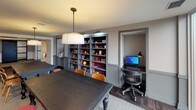 This thoughtfully designed single-floor space blends refined aesthetics with functional versatility across several interconnected zones. Dark hardwood flooring runs throughout, providing a grounding contrast to the warm gray wall tones and crisp white wainscoting that define the interior's character.

A dedicated lounge area features a tufted charcoal sofa beneath a striking starburst chandelier set within a recessed ceiling detail, creating an intimate yet dramatic focal point. Adjacent seating includes cognac leather armchairs paired with sleek pedestal side tables, alongside framed wall art that adds a curated, gallery-like quality to the space.

The layout opens into a more expansive area anchored by a large dark wood table surrounded by mixed seating. Built-in shelving units with gray-blue cabinetry line the walls, displaying an organized collection of books and decorative objects, while a dedicated computer workstation is neatly recessed into an alcove nearby.

A corridor with a plaid upholstered bench provides additional transitional space, connecting the zones fluidly. Throughout, layered lighting—including pendant fixtures, adjustable task lamps, and recessed ceiling lights—ensures each area is well-illuminated and purposefully lit, contributing to the space's polished, sophisticated ambiance.