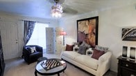 This well-appointed single-floor space showcases a thoughtfully designed living room that balances comfort and style. The room centers around a plush white sofa adorned with an array of decorative pillows in rich tones of burgundy, gray, and black floral patterns, creating a sophisticated yet welcoming atmosphere. A striking large-scale abstract artwork in warm earth tones serves as a bold focal point on the wall, adding an artistic touch to the space. A sleek glass-topped coffee table, featuring a decorative chess set, anchors the seating area and adds an element of intellectual charm. Complementing the main seating is a classic black leather chair, offering additional comfort. A decorative folding screen adds depth and visual interest, while an elegant floor lamp casts a warm, ambient glow. The room benefits from natural light filtering through a window dressed with bold patterned curtains. Crown molding frames the ceiling, which features a ceiling fan with a light fixture, adding both function and a finishing architectural detail. Soft carpeting underfoot enhances the overall sense of comfort throughout the space.