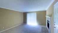 This single-floor space presents a straightforward and functional layout. The main living area features wall-to-wall carpeting in a neutral tone, complemented by warm beige walls and crisp white baseboards and crown molding that add a clean, finished look throughout. A textured popcorn ceiling adds depth overhead.

A prominent sliding glass door serves as the room's focal point, dressed with vertical blinds that allow for adjustable light control. When fully illuminated, the door floods the space with an abundance of natural light, brightening the interior considerably. The transition from carpet to tile near the door suggests an entryway or patio access point, adding a practical element to the layout.

The walls are neutral and move-in ready, providing a blank canvas for personalization. Electrical outlets are positioned conveniently along the walls, and the overall condition of the space appears clean and well-maintained. The open, unobstructed floor plan allows for flexible furniture arrangement, making it adaptable to a variety of living configurations. The combination of natural light, neutral finishes, and simple architectural details creates a calm, livable environment.