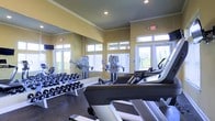This single-floor fitness center is designed to support a comprehensive workout experience. The space is outfitted with a row of treadmills and cardio machines positioned near large, multi-pane windows that allow abundant natural light to flow in, offering views of the outdoors and contributing to an open, airy atmosphere. Crown molding lines the upper walls, adding a refined architectural detail to the room.

A full rack of dumbbells in graduated weights is arranged along one wall, providing ample options for strength training. A large floor-to-ceiling mirror spans the same wall, visually expanding the space and allowing users to monitor their form during exercise. The warm yellow-toned walls contribute to an energizing ambiance throughout.

Recessed lighting in the ceiling supplements the natural light, ensuring the space remains well-lit at all times. Safety is addressed with clearly marked emergency exit signs. The durable flooring is suited for the demands of a fitness environment. Overall, the space is thoughtfully arranged to balance cardio, strength, and functional training needs efficiently.