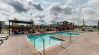 This outdoor amenity space is situated within a contemporary multi-story building complex, combining modern architectural elements such as mixed brick, concrete block, and fiber cement panel cladding in neutral tones accented by slate blue siding. The courtyard is thoughtfully organized to accommodate a variety of recreational and leisure activities.

At its center, a generously sized swimming pool with clear turquoise water serves as the focal point, complemented by an adjoining raised spa or wading area. Stainless steel entry rails provide safe pool access, and the surrounding concrete deck offers ample space for movement and relaxation. A row of white sling-style chaise lounges lines the sun deck along the building's base, positioned to capture open sky and natural light throughout the day.

A dedicated activity zone features a full-sized ping-pong table alongside bar-height seating and large ornamental planters with seasonal foliage, adding color and texture to the hardscape. A covered pergola-style structure with additional seating and teal market umbrellas offers a shaded gathering area toward the perimeter. Large glass doors provide direct interior access to the building, seamlessly connecting indoor and outdoor spaces. The elevated balconies of the surrounding structure overlook the courtyard, reinforcing the sense of an integrated, community-oriented environment.