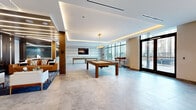 Located at 329 Alfred Ave in Teaneck, NJ, this contemporary amenity lounge is a well-appointed, single-floor recreational space offering a sophisticated and inviting atmosphere. The open-plan layout is anchored by large-format stone-look tile flooring and a tray ceiling with integrated LED cove lighting that lends a warm, modern glow throughout.

The main area serves as a game and social hub, highlighted by a solid wood pool table at its center and a shuffleboard table positioned near the rear. A wall-mounted flat-screen TV backed by a textured horizontal-stripe accent wall adds an entertainment focal point. Adjacent to the game area, a comfortable lounge zone features plush seating arranged over a patterned area rug, creating a relaxed gathering space.

Tucked to one side is an intimate private meeting nook with curved bench seating, a round table, and decorative pendant lighting — ideal for small group conversations or informal meetings. A glass-enclosed corridor connects to additional building areas, offering seamless flow.

Floor-to-ceiling windows along one full wall flood the space with abundant natural light and frame views of the surrounding courtyard and community. Direct access to an outdoor terrace further extends the social possibilities. Rich wood column accents and gold-toned fixtures complete the polished, upscale design aesthetic of this versatile amenity space.