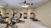 The Cedar Creek Circle Fitness Center in Montgomery, Alabama, is a single-floor community fitness facility designed to provide residents with a convenient and functional space for their exercise needs. The main workout area features wall-to-wall carpeting and is equipped with a selection of cardio machines, including two treadmills, an elliptical cross-trainer, and a stationary bike. A large wall mirror enhances the sense of space and allows users to monitor their form during workouts. Overhead, a ceiling fan with an integrated light fixture provides air circulation and illumination, complemented by recessed lighting and a fluorescent fixture. The warm, neutral-toned walls create a clean and comfortable environment. Adjacent to the main exercise room is a welcoming lounge or reception area, featuring tile and hardwood flooring, a decorative area rug, and comfortable seating including a sofa and accent chair. Natural light filters in through large windows fitted with blinds, and a glass-paneled entry door opens to the outdoors. Wall art and a side table add a homey touch to this transitional space, making it an inviting entry point into the fitness center.