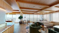 Located in Hollywood at 6390 De Longpre Ave, this elegantly designed lounge space occupies a single floor with sweeping panoramic views of the Los Angeles skyline. The open-plan layout is unified by herringbone-patterned hardwood flooring and a striking ceiling defined by warm wood beams and recessed lighting, creating a refined yet welcoming atmosphere throughout.

The main lounge area is anchored by a generous curved sectional sofa in soft neutral tones, complemented by bold emerald green velvet club chairs and organic-shaped coffee tables in brass and wood. A curated patterned area rug ties the seating areas together, while a large flat-screen display and decorative credenza add a media and entertainment dimension to the space.

A standout feature is the curved wet bar, finished with fluted light wood cabinetry, a veined marble countertop, a brass detail rail, and white tile backsplash accented by integrated under-cabinet lighting — a centerpiece of refined craftsmanship. Adjacent to the main lounge, a cozy dining nook offers an intimate seating area with a round marble-top table, colorful accent chairs, and original artwork on the wall.

Floor-to-ceiling windows with adjustable venetian blinds and sheer drapes flood the entire space with natural light while framing unobstructed city views, enhancing the elevated ambiance of this thoughtfully appointed space.