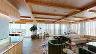 Located in Hollywood at 6390 De Longpre Ave, this elegantly designed lounge space occupies a single floor with sweeping panoramic views of the Los Angeles skyline. The open-plan layout is unified by herringbone-patterned hardwood flooring and a striking ceiling defined by warm wood beams and recessed lighting, creating a refined yet welcoming atmosphere throughout.

The main lounge area is anchored by a generous curved sectional sofa in soft neutral tones, complemented by bold emerald green velvet club chairs and organic-shaped coffee tables in brass and wood. A curated patterned area rug ties the seating areas together, while a large flat-screen display and decorative credenza add a media and entertainment dimension to the space.

A standout feature is the curved wet bar, finished with fluted light wood cabinetry, a veined marble countertop, a brass detail rail, and white tile backsplash accented by integrated under-cabinet lighting — a centerpiece of refined craftsmanship. Adjacent to the main lounge, a cozy dining nook offers an intimate seating area with a round marble-top table, colorful accent chairs, and original artwork on the wall.

Floor-to-ceiling windows with adjustable venetian blinds and sheer drapes flood the entire space with natural light while framing unobstructed city views, enhancing the elevated ambiance of this thoughtfully appointed space.