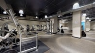 This expansive fitness center is designed to support a wide range of workout preferences across several distinct zones. The main gym area features durable speckled rubber flooring throughout, a dark exposed ceiling with visible ductwork and pendant lighting, and large wall-mounted mirrors that enhance the sense of space. Cardio equipment, including multiple treadmills, ellipticals, and stationary bikes, is spread across dedicated sections, providing ample room between machines. A free weights area offers a full dumbbell rack alongside mirrored walls for proper form reference.

A separate studio space is a standout feature, boasting polished light hardwood floors, acoustic wall panels, a wall-mounted flat-screen TV, and interior windows overlooking the main gym. Wall-mounted ballet barres and yoga mats make it well-suited for yoga, stretching, or group fitness classes. The studio is illuminated by pendant lights and benefits from additional natural light through interior-facing windows.

An open recreation area rounds out the facility, offering ping-pong tables, a punching bag, and medicine ball storage, all set against the same industrial-style dark ceiling and rubber flooring. The overall ambiance is clean, functional, and modern.