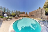 Located at 725 College Ave in Santa Rosa, California, this single-level outdoor space centers around a generously sized freeform swimming pool with a distinctive kidney-shaped design. The pool's clear turquoise water is complemented by a smooth concrete pool deck that wraps around the perimeter, providing ample space for lounging and movement. The poolside area is well-furnished with multiple seating arrangements, including tables and chairs shaded by large market umbrellas, as well as additional lounge chairs for sunbathing. Restroom and utility facilities are conveniently accessible along one edge of the pool area. The space is enclosed by cedar shake-clad walls and wood fencing, lending a warm, natural aesthetic that harmonizes with the surrounding landscape. Mature trees, including tall conifers and leafy deciduous varieties, frame the area and provide a natural canopy overhead, enhancing privacy and shade. Manicured hedgerows and planted greenery line the walkways, adding to the lush, park-like atmosphere. The overall ambiance is tranquil and well-maintained, offering a comfortable outdoor retreat suited for relaxation and recreation in the mild Northern California climate.