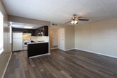 Located in El Paso, TX, this single-floor space offers a practical and functional layout with consistent design elements throughout. The main living area is highlighted by wood-look plank flooring that extends across the space, a textured popcorn ceiling, and a ceiling fan with integrated lighting that provides both airflow and illumination. A distinctive vertical wood panel accent wall adds a subtle architectural detail to the living room, complementing the neutral, warm-toned walls throughout.

The kitchen features dark espresso-toned cabinetry with raised panel detailing on both upper and lower cabinets, paired with light-colored countertops that offer a visual contrast. Appliances include a white electric range, a white refrigerator, and a stainless steel dishwasher. An overhead range hood is also present. The kitchen opens toward the dining area through a pass-through counter arrangement, allowing for a connected flow between spaces. The dining area benefits from natural light provided by multiple windows with blinds.

The bedroom area continues the wood-look flooring and features louvered closet doors for storage. A floor vent and wall-mounted air register indicate a central HVAC system. The textured ceilings and neutral color palette are consistent throughout, providing a cohesive and understated aesthetic across the entire space.