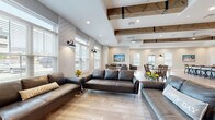 Crestwood Commons is a single-story community clubhouse located on Judge Carden Blvd in Crestwood, Kentucky, thoughtfully designed to blend modern farmhouse aesthetics with practical amenity spaces. The welcoming entry foyer greets visitors with wainscoting, a mounted television, leather seating, and decorative accents that set the tone for the entire facility.

The main gathering area is expansive and open, featuring exposed wood beam ceiling details, shiplap accent walls, and large windows that bathe the space in natural light. Durable wood-look tile flooring flows throughout, unifying the lounge seating area—furnished with leather sofas—and a dedicated dining zone complete with wood tables and bench seating.

The full kitchen is well-appointed with dark cabinetry, granite countertops, a subway tile backsplash, stainless steel appliances, and an oversized island with industrial-style bar seating. A convenient beverage and snack station with granite counters and a coffee maker provides additional functionality for residents.

The fitness center makes a bold statement with vibrant red and blue accent walls, rubber flooring, cardio equipment, weight benches, storage lockers, and a ceiling fan for airflow. A tanning bed unit is also available within the facility. Throughout the space, equestrian-themed artwork adds a nod to Kentucky's rich horse country heritage.