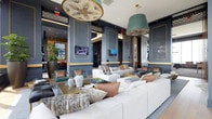 This stylish lounge space makes a striking first impression with its sophisticated design palette. The walls are finished in a deep slate blue, accented with gold trim that frames paneled sections, adding a sense of refined structure and visual depth throughout the room. The ceiling draws the eye upward, where two statement light fixtures anchor the space — a large medallion-style pendant with an ornate floral design and a bold teal drum shade pendant, both contributing dramatically to the room's upscale character.

The seating area is generously appointed with oversized white sofas and loveseats, layered with an eclectic mix of throw pillows in warm tones of copper, blush, teal, and taupe. Acrylic side tables and low-profile coffee tables provide functional surfaces while maintaining a light, modern aesthetic. Multiple flat-screen televisions are mounted on the walls, enhancing the space's utility as a social and entertainment hub.

Lush greenery, including a tall potted plant in a dark planter, introduces a natural element that softens the polished décor. Light wood flooring ties the design together, offering warmth against the cooler wall tones. Overall, the space exudes a contemporary yet welcoming ambiance ideal for social gatherings.